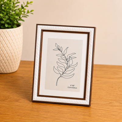 Porta retrato amadeirado e branco com ilustração de folha em fundo claro, ao lado de vaso com planta em ambiente de madeira